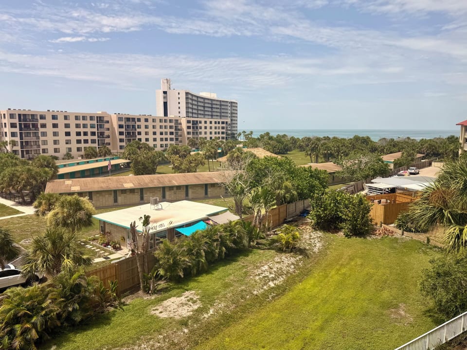 The property sits in a tranquil coastal setting with lush landscaping, tropical vegetation, and neighboring beachfront buildings visible in the distance. The Gulf waters provide a serene backdrop to this relaxed vacation destination.