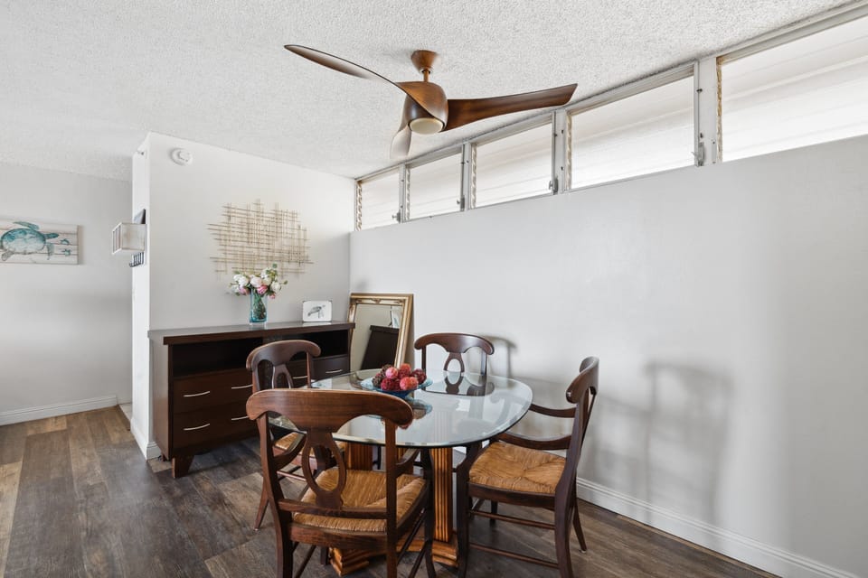 Dining nook with glass‑top table and fresh flowers, a breezy spot to gather under the wooden‑bladed fan.