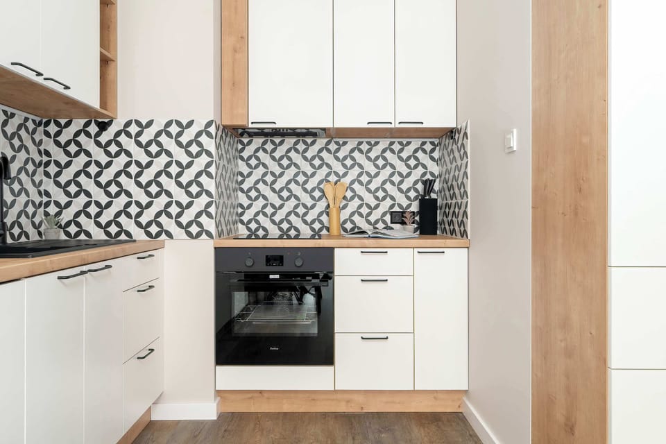 A compact contemporary kitchen with upper and lower cabinets and a geometric backsplash. The design blends wood and white surfaces for a fresh, clean look.