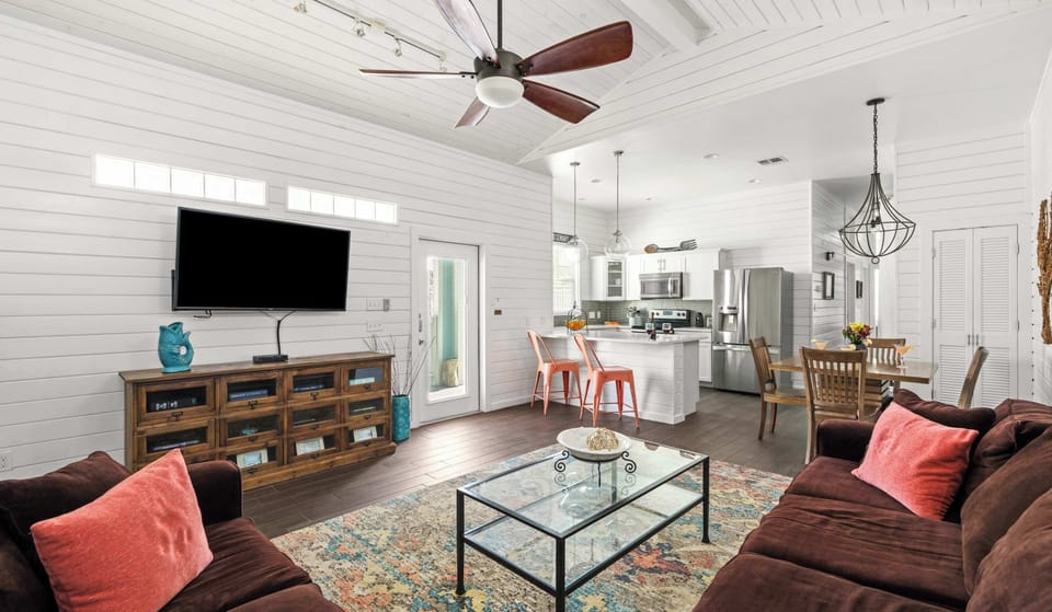 Living Room & Kitchen — Brown Sectional, Coral Pillows, Glass Coffee Table, Colorful Rug, Wall TV & White Island