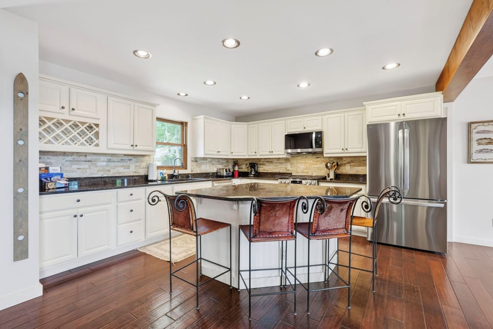 A large white kitchen with dark wood floors and an eat - at island. A large stainless refrigerator with two doors and a lower freezer. A wine rack above the dark counter.