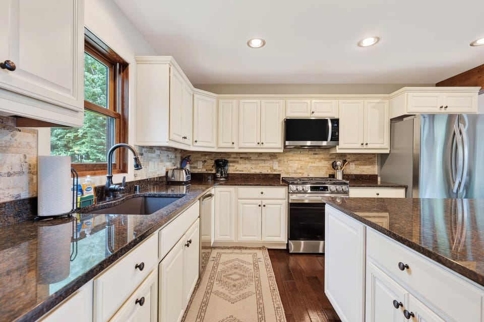 A large white kitchen with dark wood floors with a sink with a tall faucet. Gas Stove a microwave above it.