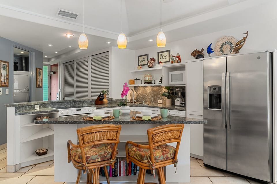 A bright kitchen featuring a granite island with bar seating, pendant lighting, and stainless steel appliances