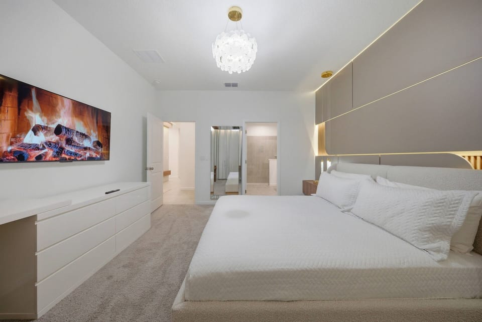 Relax in a serene bedroom adorned with soft neutral tones. The spacious layout features a comfy bed, elegant nightstands, and a modern entertainment unit, creating the perfect ambiance for cozy evenings under a stylish chandelier.
