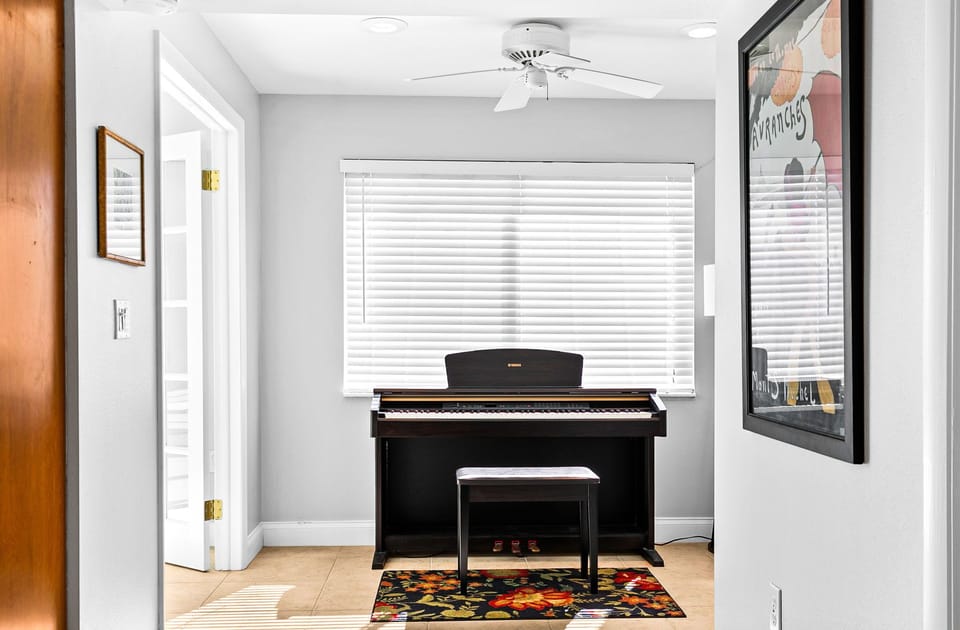 Step into your creative sanctuary where a beautiful piano awaits, positioned perfectly beneath bright windows. The soft ceiling fan keeps you comfortable as you play, while colorful artwork adds warmth to this inspiring musical corner of your home.