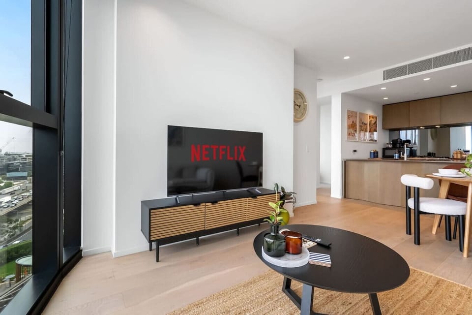 Open-plan living space with a large smart TV, city views to the left, and a fully equipped kitchen visible in the background. Clean lines, warm timber finishes, and a dining area make it a comfortable space for both relaxing and entertaining.