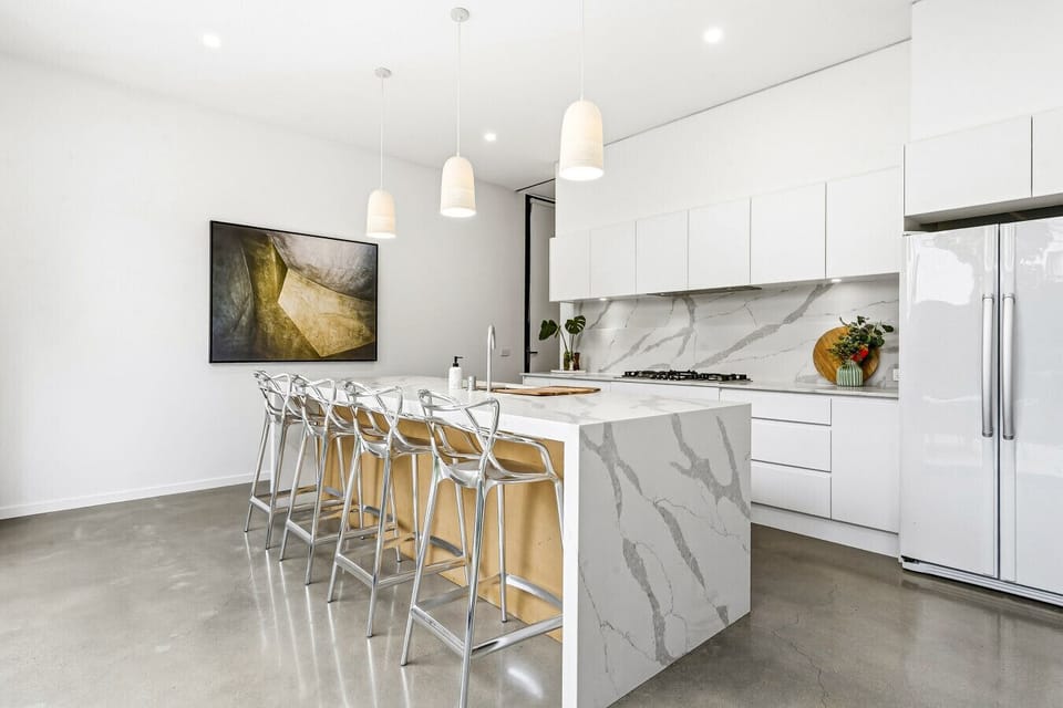Modern kitchen with a marble waterfall island and breakfast bar seating for four, gas cooktop, and full-height splashback. Pendant lighting, a side-by-side fridge, and polished concrete floors complete the space.