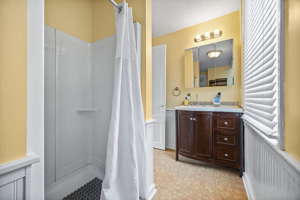 A bathroom with yellow walls, white wainscoting, a dark wood vanity, and a shower.