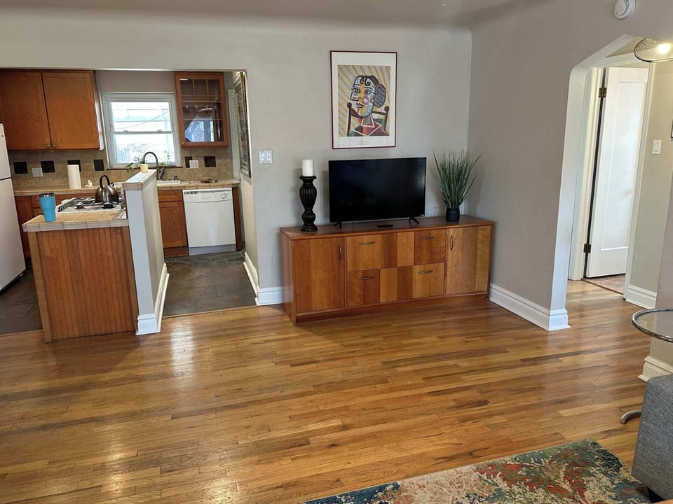 Living room detail featuring a mid-century modern wooden sideboard used as a TV stand. Hardwood floors and neutral walls create a versatile backdrop for decor, with a glimpse into the kitchen area.