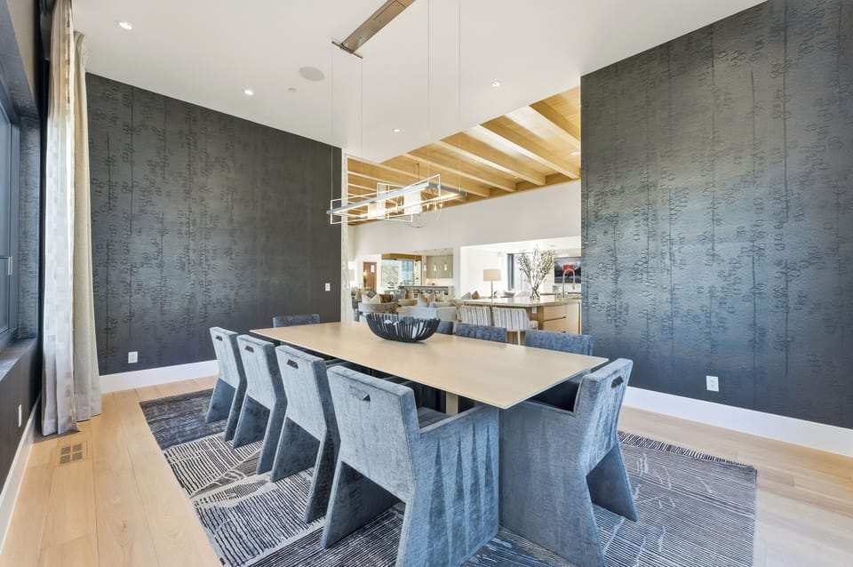 Spacious dining room open to kitchen, featuring vaulted ceilings, contemporary lighting, and black upholstered chairs.