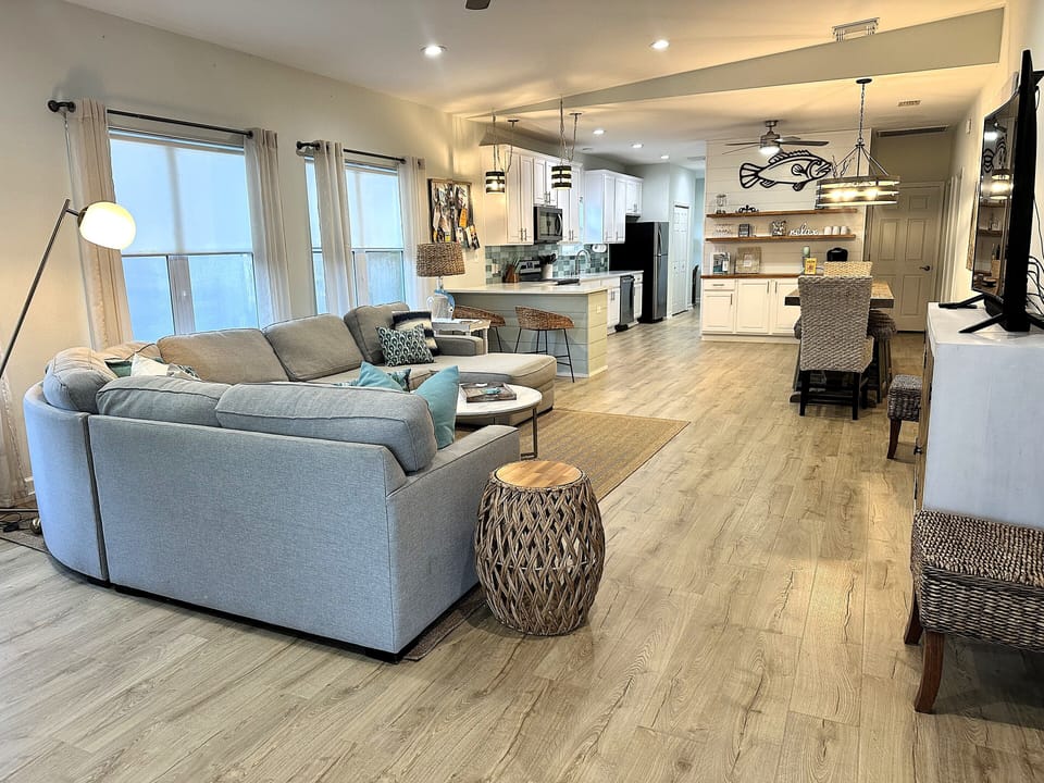 Cozy beach vibes with a gray sectional, white kitchen, woven chairs, & fish art.