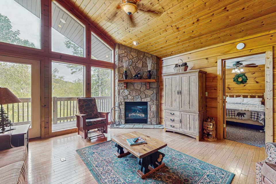 Bright cabin living space with stone fireplace, wall of windows, and peaceful mountain views. Perfect for cozy mornings and relaxing evenings. 