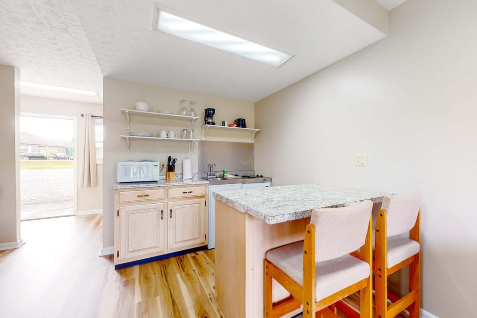 Compact kitchenette with breakfast bar seating. Perfect for quick meals, coffee, and long stays. Studio-friendly suite with mini kitchen, microwave, sink, dishes, and convenient dining space.