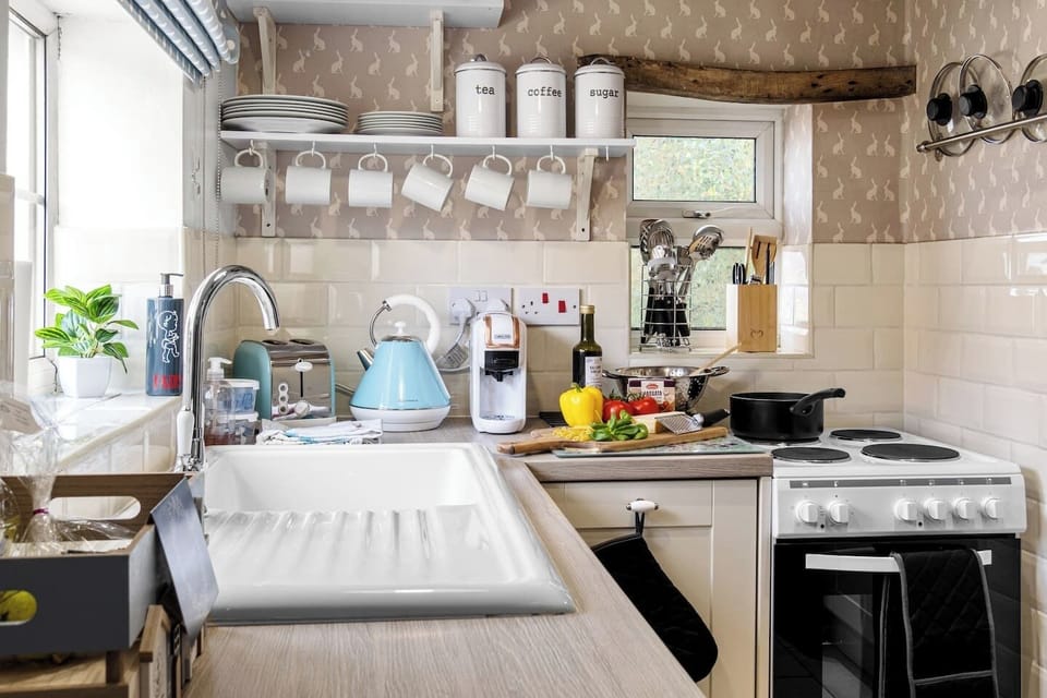 A wide kitchen worktop view with sink, mugs, shelving, and cooker.