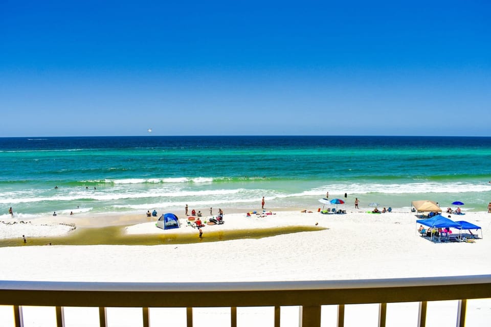 From the balcony, beachgoers can be spotted enjoying the lively atmosphere below, with colorful umbrellas dotting the shoreline as gentle waves create a captivating backdrop to a perfect day by the ocean.