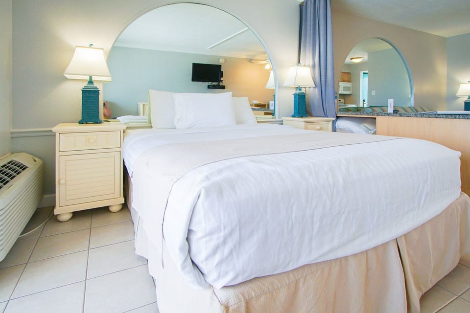 The cozy bedroom features a comfortable bed draped in crisp white linens, complemented by charming ocean-blue lamps and a mirrored wall that amplifies the inviting atmosphere of this beach-side retreat.