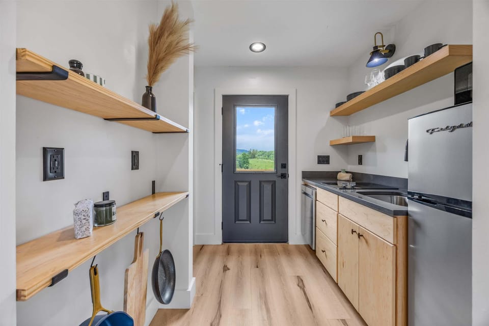 Bright galley kitchen with open shelving, modern appliances, and a door framing scenic outdoor views.