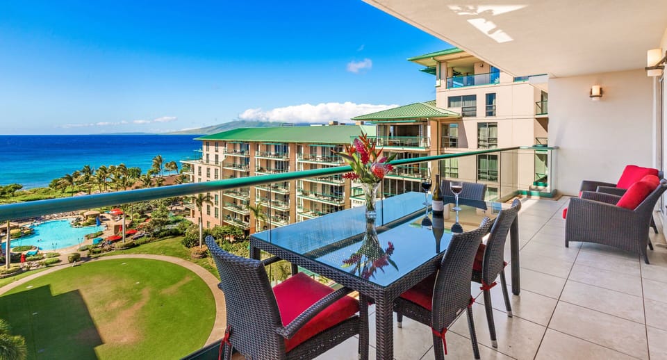 Ocean-view balcony with a glass-top dining table and wicker chairs accented by red cushions, overlooking resort grounds and turquoise waters.