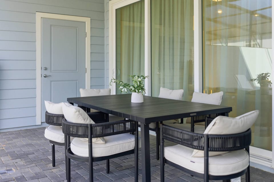 Enjoy the charming patio with a sleek black table and cushioned chairs, perfect for morning coffee or evening meals. The soft furnishings complement the light siding and large glass doors, inviting you to unwind in comfort.
