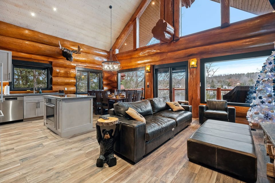 Bright and inviting main living area featuring a large sectional sofa, handcrafted log beams, and stunning views overlooking the lake.