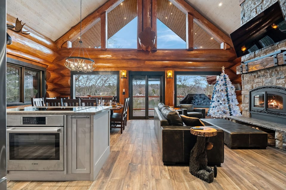 Spacious open-concept great room with vaulted ceilings, wall-to-wall windows, cozy leather seating, and a real wood-burning fireplace—perfect for family gatherings.