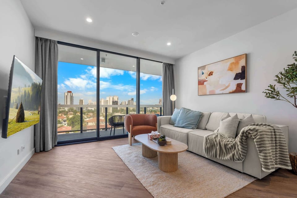 A bright and open lounge space styled with warm timber tones and curved accents, flowing seamlessly onto the balcony overlooking the city.