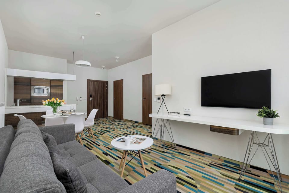 A contemporary living room containing a gray sectional sofa, mounted TV, and a dining table set on a colorful patterned rug.