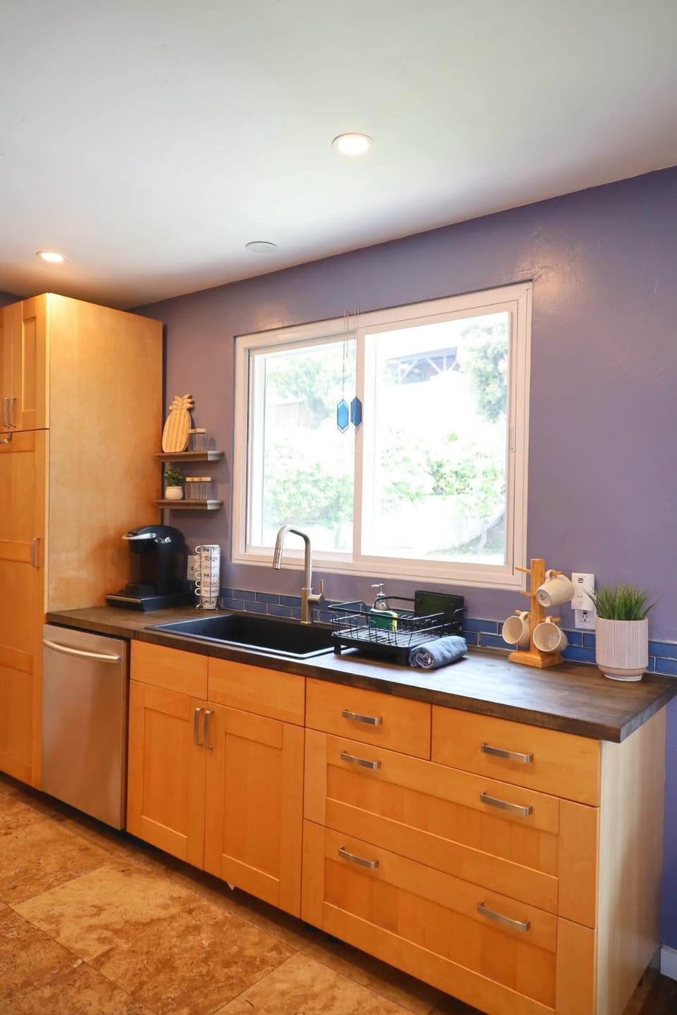 A bright kitchen with light wood cabinets, a dark counter, and a coffee maker and a few cozy touches.