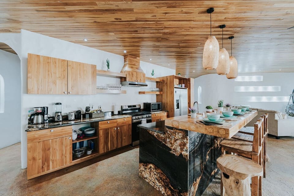 This open kitchen feels warm and modern with its teak ceiling, stone counters, stainless appliances, and rich wooden cabinetry. The generous eat-in island makes it an easy place to cook, gather, and spend time together.