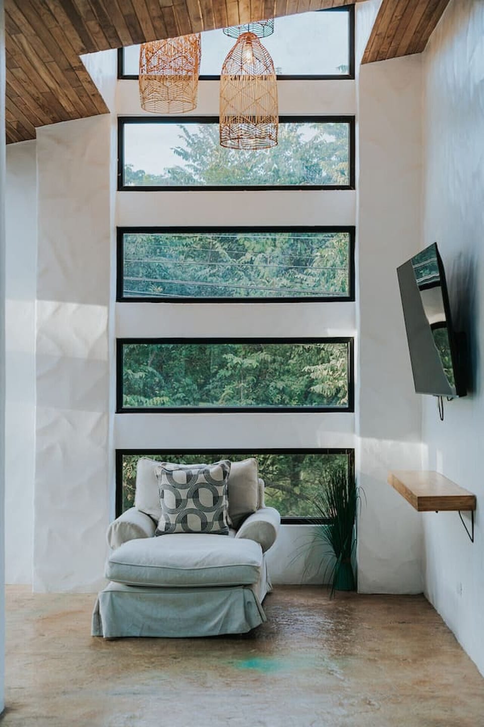 Stretch out on the club sofa in the upstairs living area. Floor-to-ceiling windows frame lush greenery, while white textured walls, high ceilings, rattan lights, and a smart TV create a bright, stylish space.