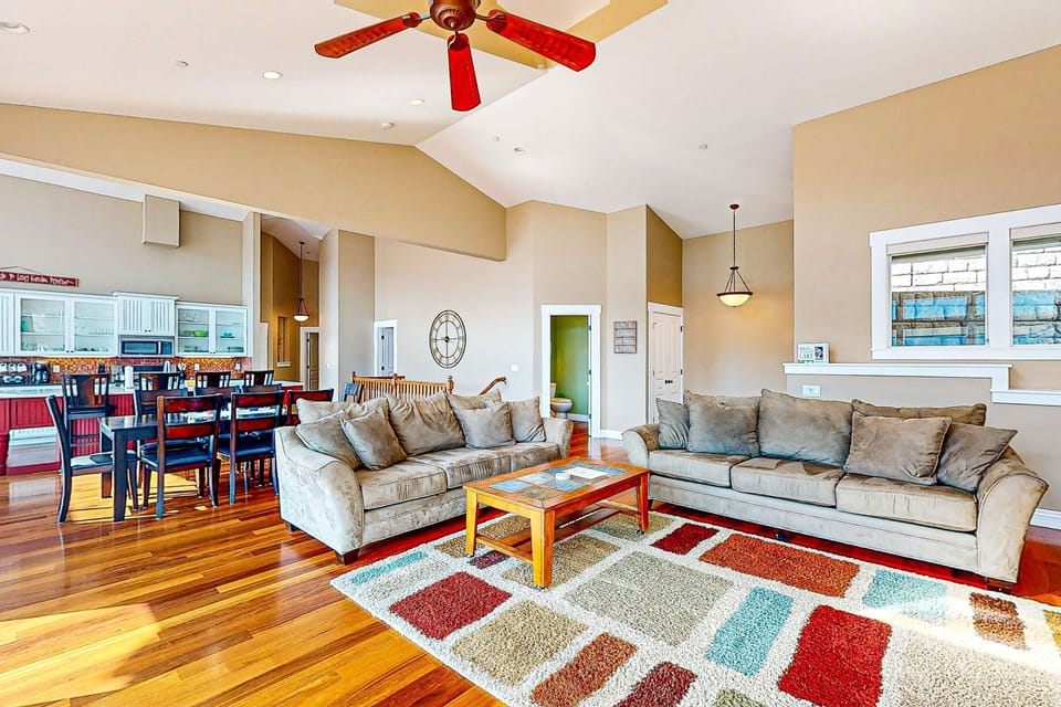 Open-plan living with vaulted ceilings, dual sofas, hardwood floors, and dining area open to the white-cabinet kitchen — room for the whole group