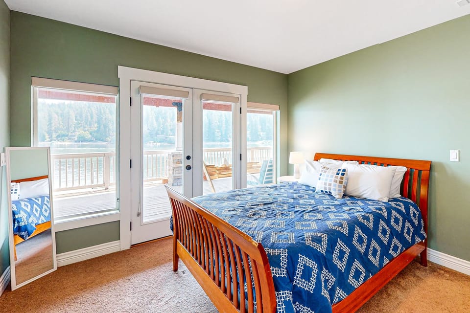 Guest bedroom with wood-frame sleigh bed, French doors opening to lake-view deck, and wooded shoreline views — fall asleep to the water