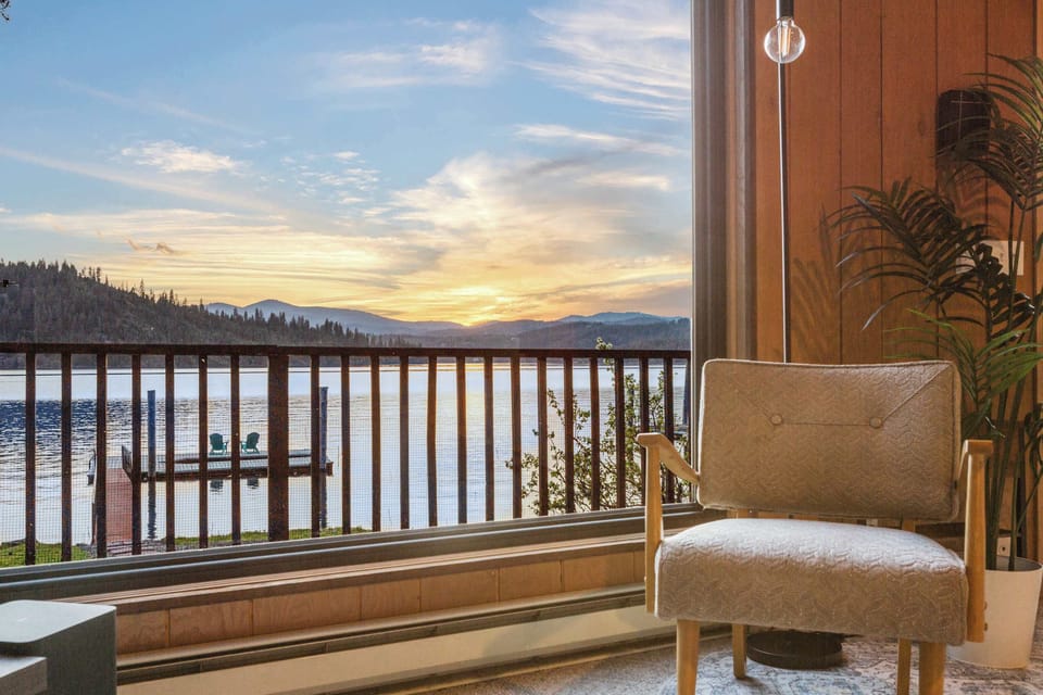 Twilight lake-view reading nook — accent armchair, floor-to-ceiling windows, and a sunset over the water that frames the dock perfectly