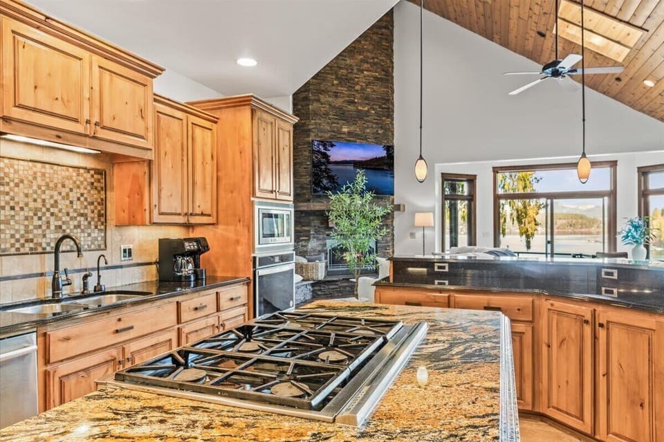 Kitchen with granite island cooktop, mosaic tile backsplash, and soaring stone fireplace wall flowing into the living area
