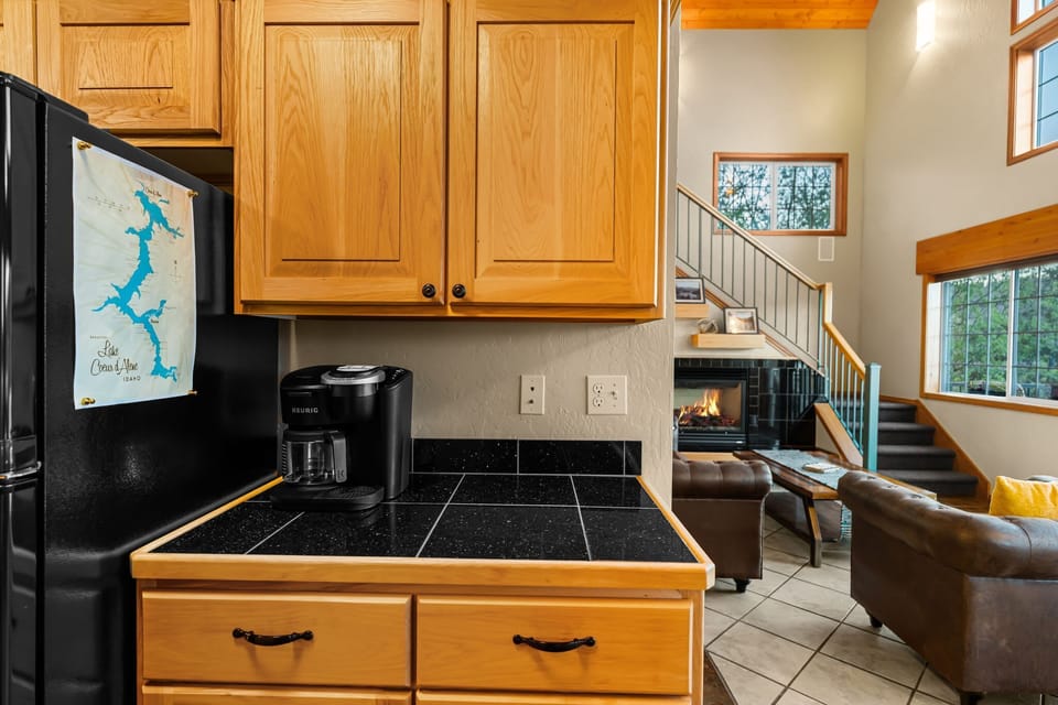 Kitchen pass-through angle with dark tile counters, Keurig station, and an open sightline to the lit fireplace and living room beyond.