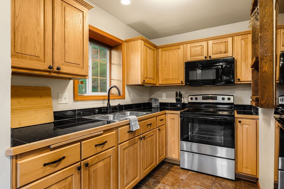Kitchen with wood cabinets, dark tile counters, double sink, stainless range, and a garden window framing the tree-lined hillside.