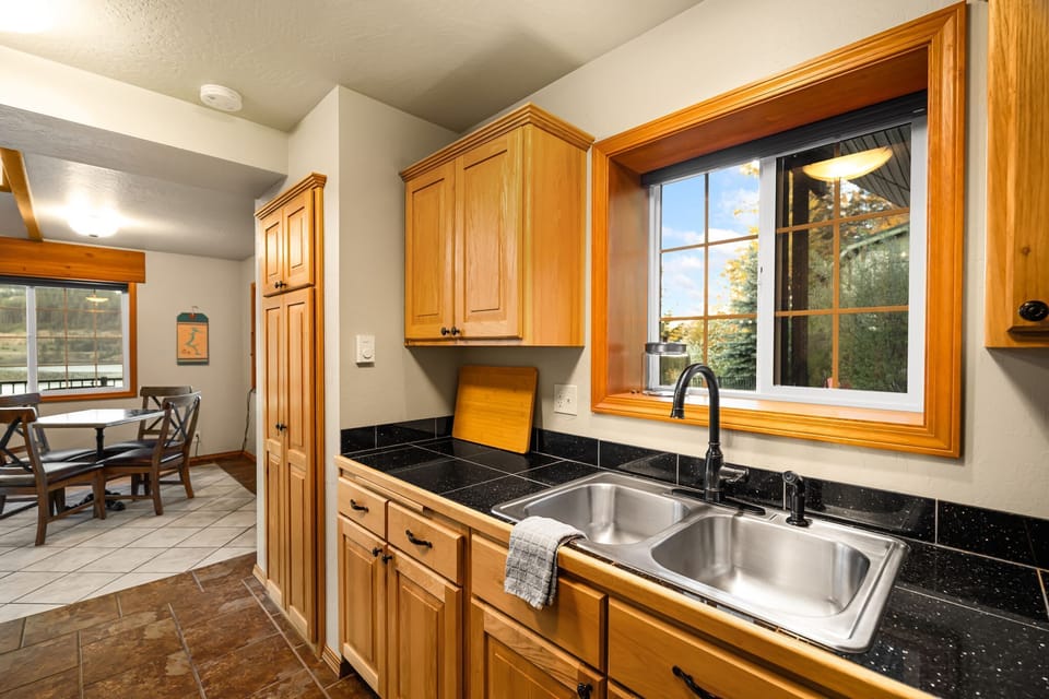 Kitchen sink area with double basin, dark tile counters, wood cabinetry, and a bright window view of the lake patio beyond.