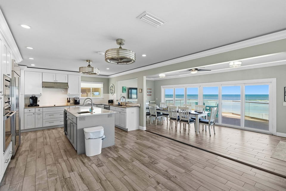 Bright open-concept kitchen and dining area with modern finishes, large island, and floor-to-ceiling sliders leading to a beachfront balcony—perfect for your beach stay with stunning ocean views and indoor-outdoor living.