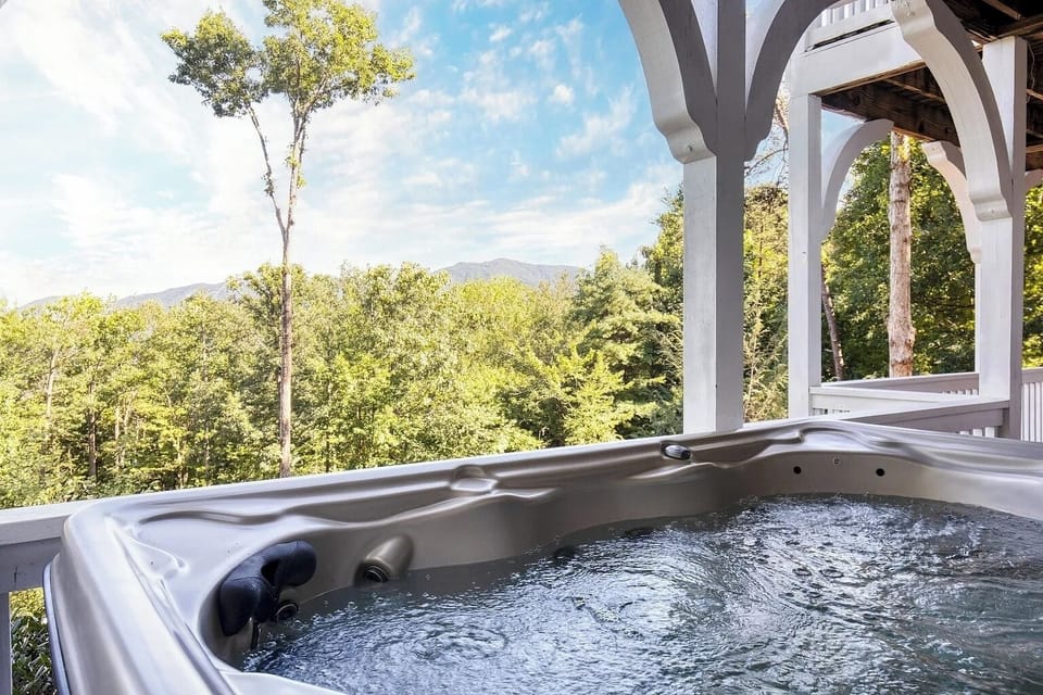 A luxurious hot tub sits beneath a charming covered deck overlooking lush forest scenery. The space features timber columns and open sky views, perfect for relaxing while immersed in nature.
