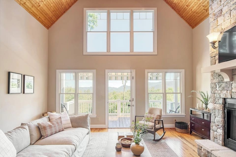 A grand living room with soaring ceilings, oversized windows, and abundant natural light. Cozy neutral furnishings, hardwood floors, and a central seating area create a warm and elegant ambiance.