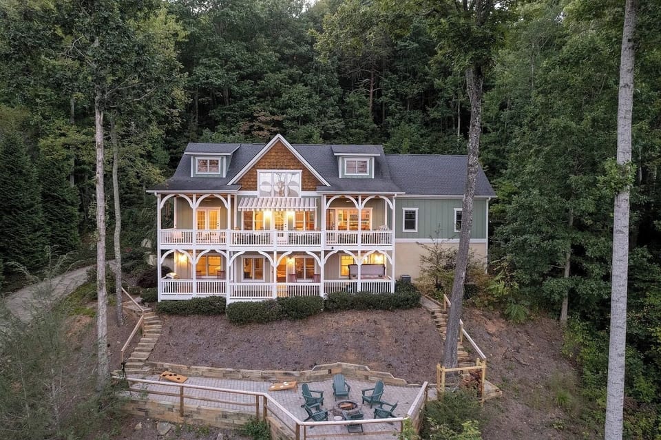 A stunning multi-level mountain home surrounded by tall pines. Warm interior lighting glows through expansive windows, and a large outdoor firepit area sits below, showcasing the property’s serene wooded setting.