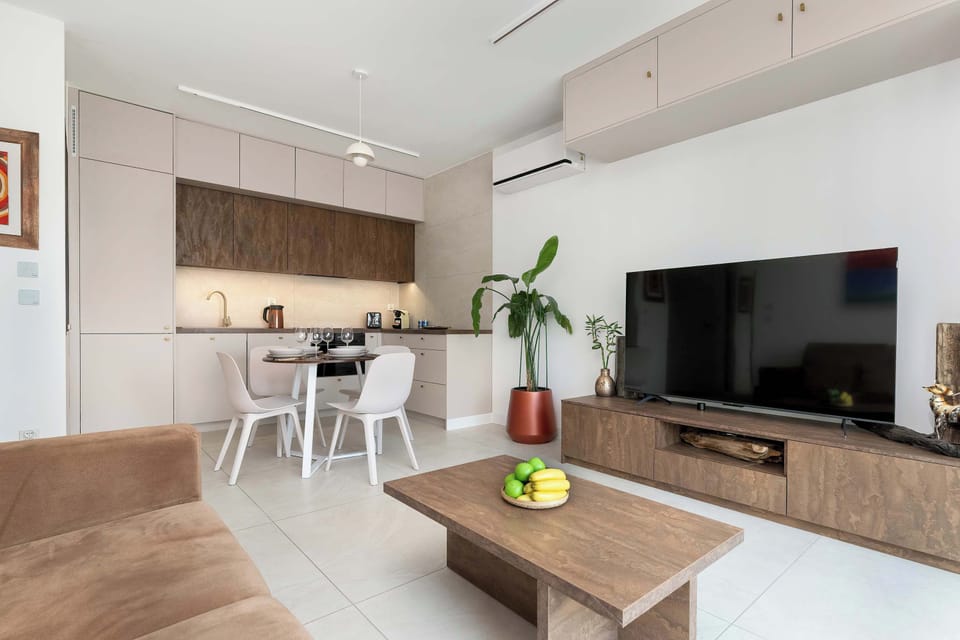 A living room corner with a sofa, colorful wall art, and a wooden coffee table. A green plant in the corner brings life into the space. The wide windows let in plenty of natural light.