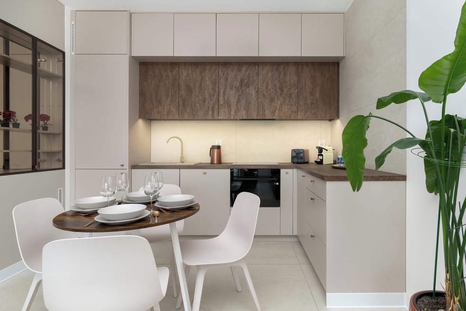 A sleek kitchen featuring white cabinetry and warm wooden accents. A round dining table with white chairs stands nearby, blending well with the minimalist design. A tall plant adds freshness to the room.