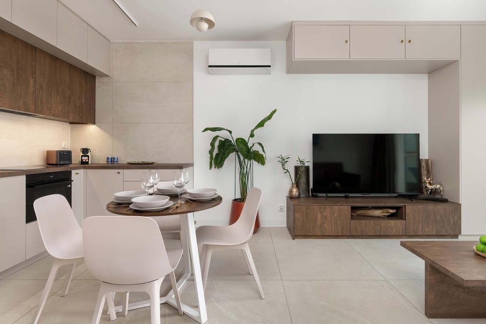 A modern open-plan living room with a small dining table beside the sofa. Bright windows illuminate the space, making the light furniture stand out. The setup feels practical and inviting.