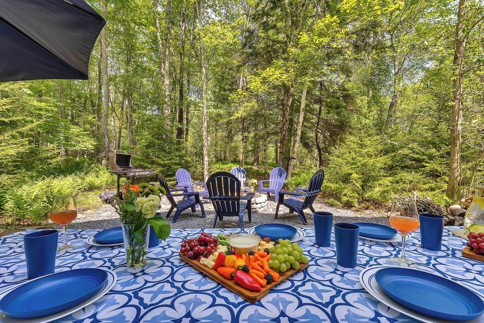 Dine alfresco in the heart of nature with this forest-side outdoor setup, complete with a vibrant picnic table, cozy fire pit seating, and a grill ready for your BBQ favorites.