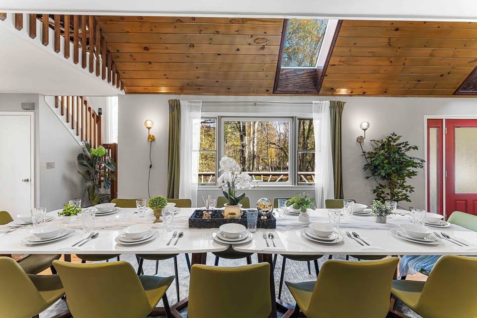 Elegant dining room with a long, beautifully set table, skylight, and large windows framing serene wooded views—perfect for memorable meals and gatherings.