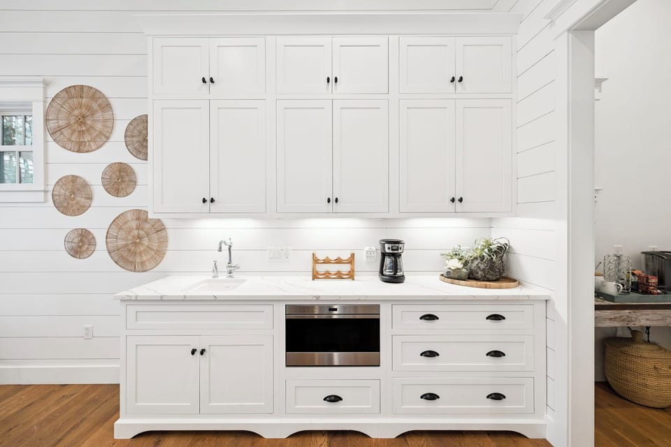 Your fully equipped kitchen blends coastal charm with modern convenience, featuring pristine white cabinetry, marble countertops, and thoughtful details like decorative woven accents that create a warm, inviting cooking space.