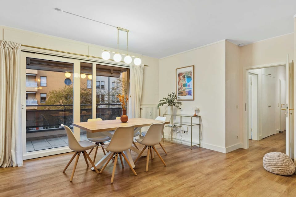 Bright open-plan living area with a cozy dining space and direct access to the balcony. Large windows fill the room with natural light, creating a warm and welcoming atmosphere for relaxing or working remotely.