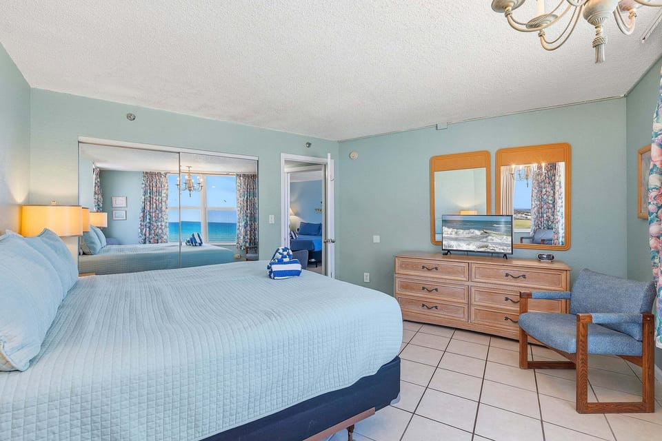 Relax in this spacious oceanfront king bedroom featuring coastal décor, a dresser with TV, mirrored closet, and stunning beach views. Designed for a comfortable beach stay with plenty of space to unwind after a day by the ocean.