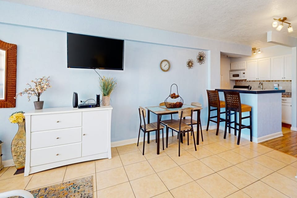 Sunny beach-style dining and living space featuring a smart TV, cozy table for four, and a breakfast bar—perfect for relaxing after a fun-filled day on the coast.